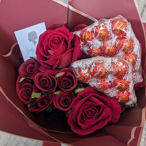 Puede incluir: Un ramo de rosas rojas y una bolsa de trufas de chocolate Lindor envueltas en papel burdeos con ribete dorado.