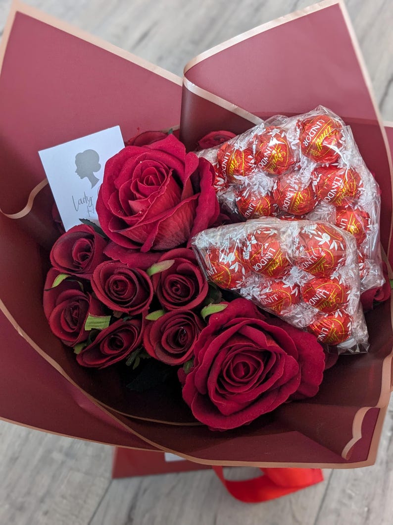Puede incluir: Un ramo de rosas rojas y una bolsa de trufas de chocolate Lindor envueltas en papel burdeos con ribete dorado.