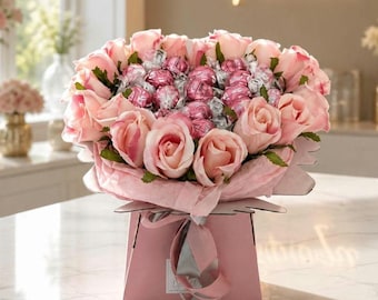 Puede incluir: Un ramo de rosas de color rosa claro intercaladas con chocolates esféricos envueltos en plata, dispuesto en una caja de regalo rosa. El arreglo está envuelto en papel de seda rosa a juego y atado con una cinta plateada, creando un diseño floral.