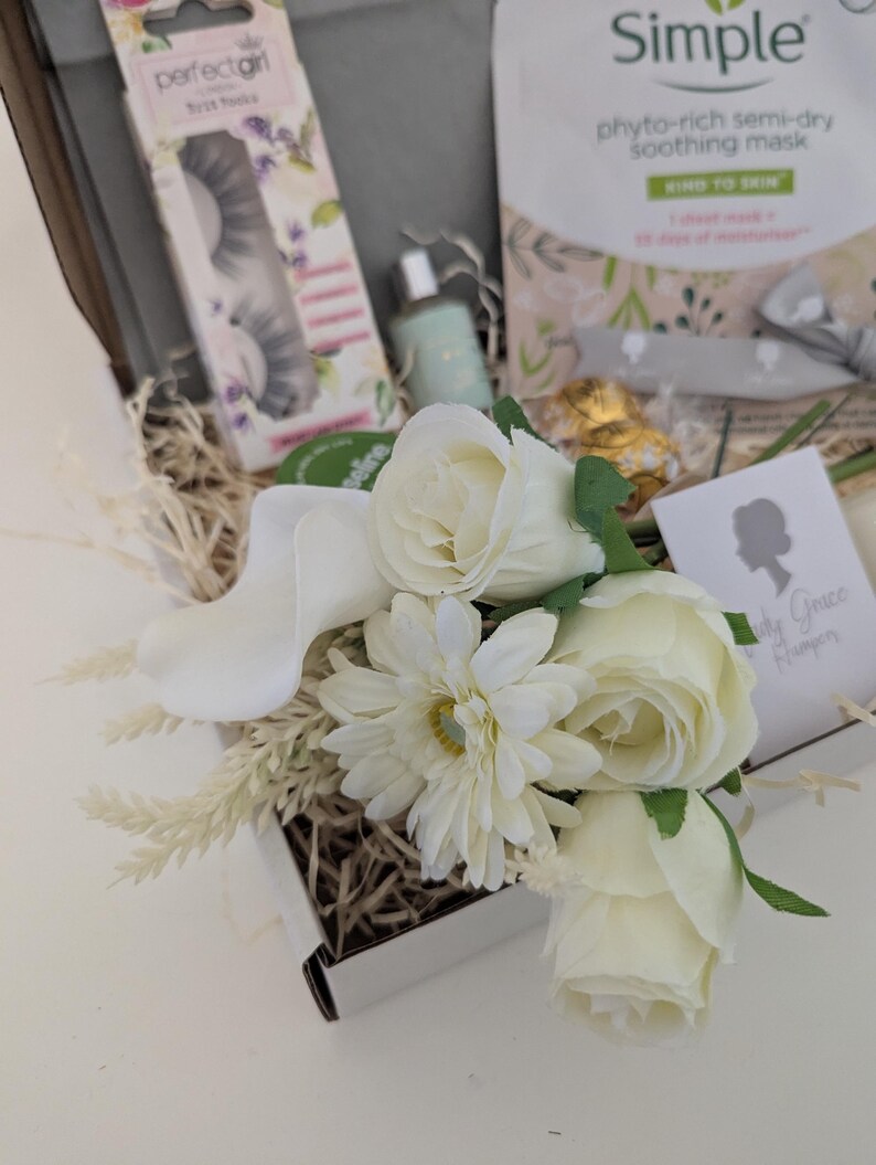 May include: A gift box filled with beauty products, including a package of false eyelashes, a face mask, and a small bottle. The box is decorated with white flowers and dried grass. The text "perfectgirl" and "Simple" are visible.