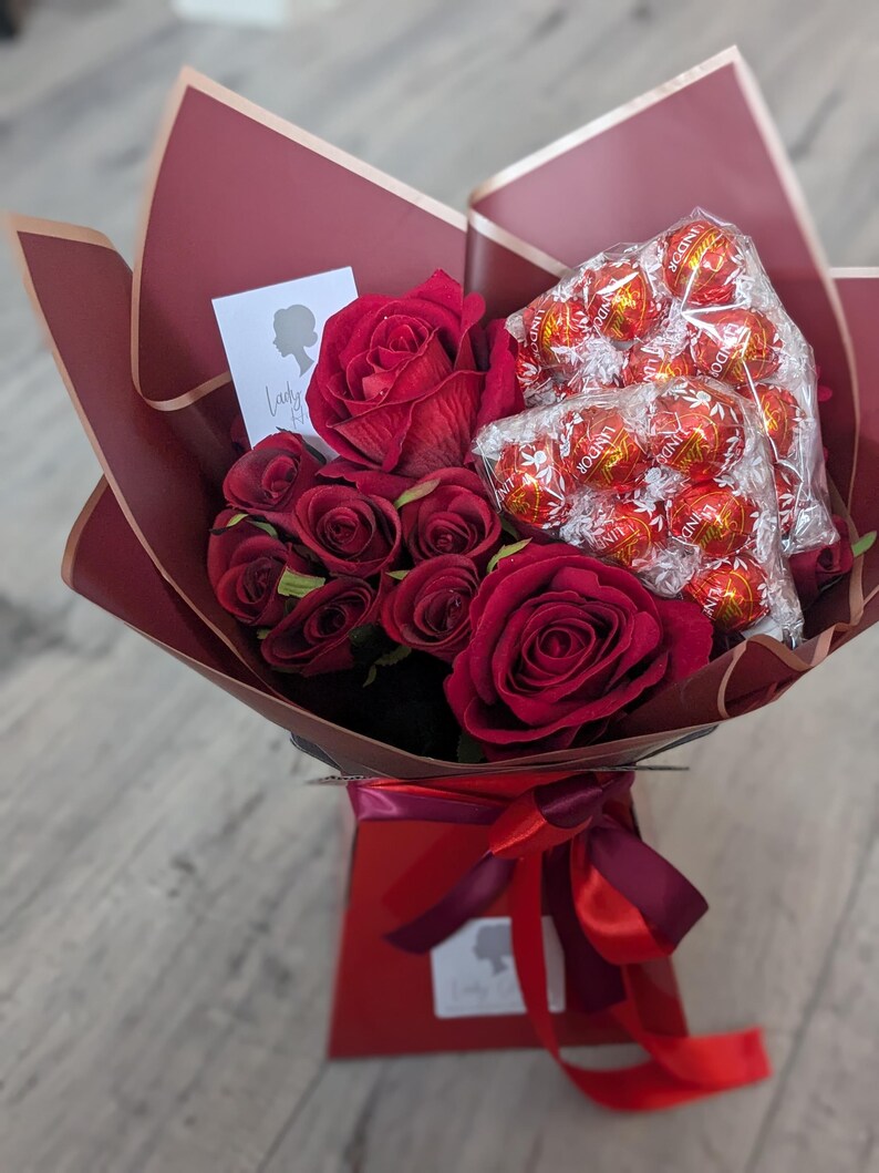 Puede incluir: Un ramo de rosas rojas y una caja de trufas de chocolate envueltas en papel burdeos con una cinta roja. El ramo est&aacute; en una caja roja con una etiqueta blanca.