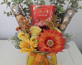 May include: A yellow gift box with a gold ribbon and a bouquet of orange and yellow flowers. The bouquet includes a box of Lindor Blood Orange chocolates.