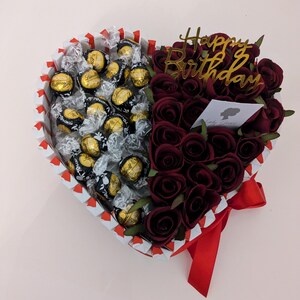 May include: A heart-shaped box filled with chocolate truffles and red roses. The box is decorated with red and white stripes and a red ribbon. The roses are arranged in a bouquet and the box has a gold "Happy Birthday" sign.