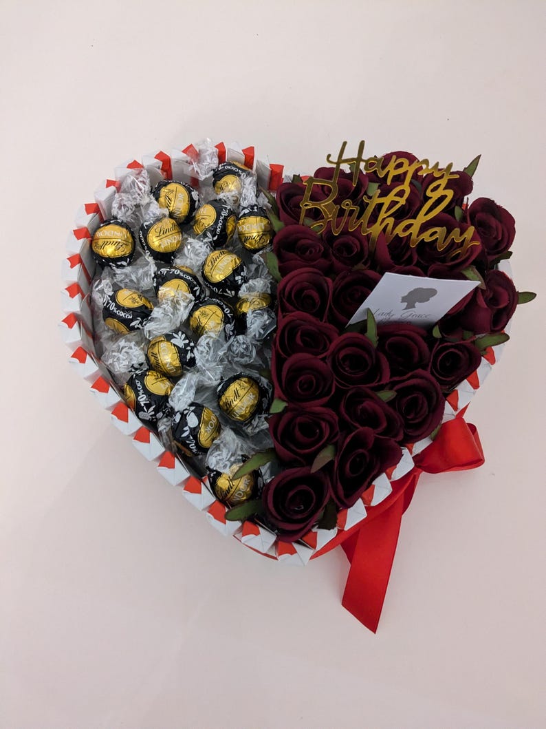 May include: A heart-shaped box filled with chocolate truffles and red roses. The box is decorated with red and white stripes and a red ribbon. The roses are arranged in a bouquet and the box has a gold "Happy Birthday" sign.