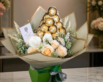 May include: A bouquet featuring a pyramid of gold-wrapped spherical chocolates, pale peach roses, white calla lilies, and a decorative butterfly. The arrangement is wrapped in light green paper, tied with a green and silver ribbon, and includes a card.
