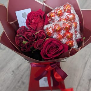 Puede incluir: Un ramo de rosas rojas y una caja de bombones Lindor envueltos en papel burdeos con una cinta roja. El ramo est&aacute; sobre una superficie de madera.