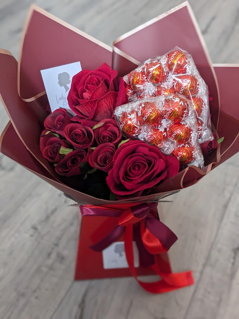 Puede incluir: Un ramo de rosas rojas y una caja de bombones Lindor envueltos en papel burdeos con una cinta roja. El ramo est&aacute; sobre una superficie de madera.