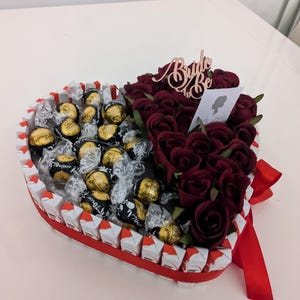 May include: A heart-shaped box filled with chocolate candies and red roses. The box is decorated with a red ribbon and a sign that says "Bride to Be".