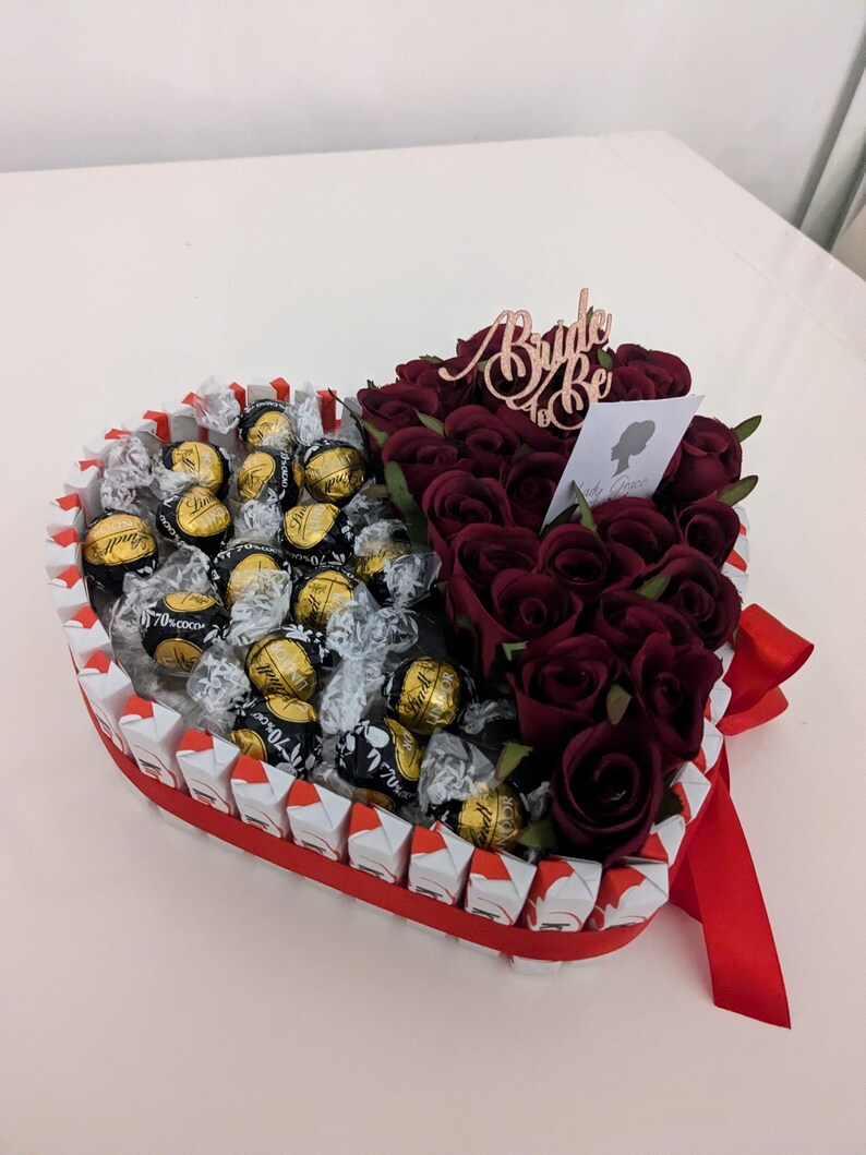 May include: A heart-shaped box filled with chocolate candies and red roses. The box is decorated with a red ribbon and a sign that says "Bride to Be".