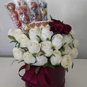 May include: A burgundy hat box filled with white roses and red roses. The box has a burgundy ribbon and the roses are arranged in a circular pattern. The box also has a small sign with the word "JESS" on it.