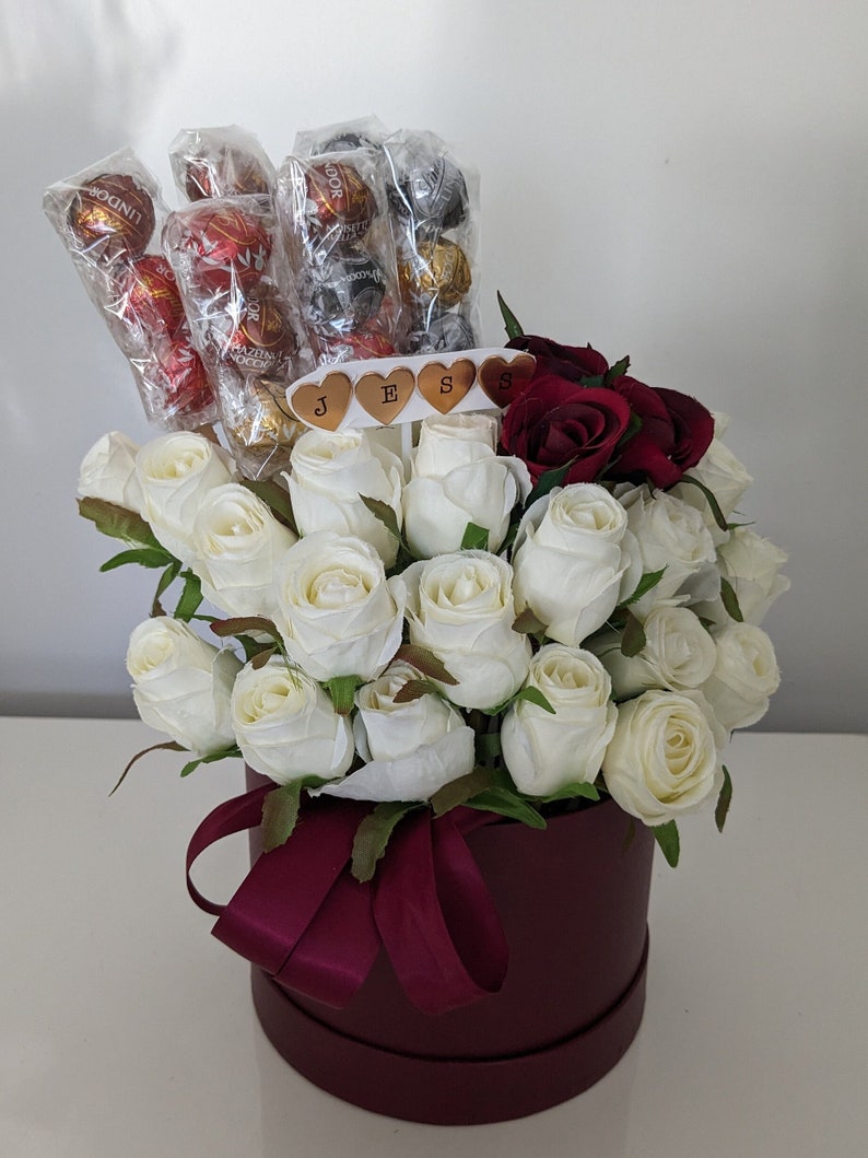 May include: A burgundy hat box filled with white roses and red roses. The box has a burgundy ribbon and the roses are arranged in a circular pattern. The box also has a small sign with the word "JESS" on it.
