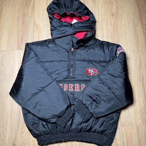 May include: Black and red hooded jacket with a half-zip closure. The jacket features the San Francisco 49ers logo on the chest and the word "49ERS" below it. The jacket has a quilted design and elastic cuffs.