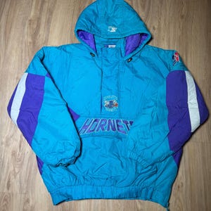 May include: A teal and purple Charlotte Hornets hooded anorak jacket. The jacket features the Hornets logo and wordmark across the front, with white stripes on the sleeves. The jacket has a drawstring hood and a Starter logo.