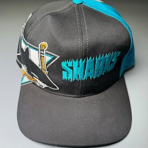 Op de afbeelding: Een zwarte en turquoise San Jose Sharks baseballpet met een haai-logo en het woord "SHARKS" geborduurd op de voorkant.