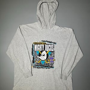 Op de afbeelding: Een grijs hoodie met lange mouwen met een grafische print van het Anaheim Mighty Ducks logo. Het logo toont een cartoon eend met een hockeymasker en twee hockeysticks. De tekst "ANAHEIM MIGHTY DUCKS" is boven het logo gedrukt.