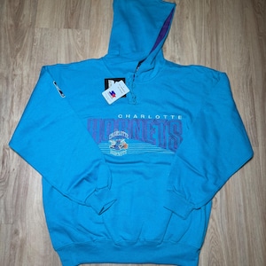 以下が含まれることがあります： 紫と白で「CHARLOTTE HORNETS」の文字が入った、ターコイズブルーのパーカー。紫の裏地とタグが付いた、ドローストリングフードが付いています。