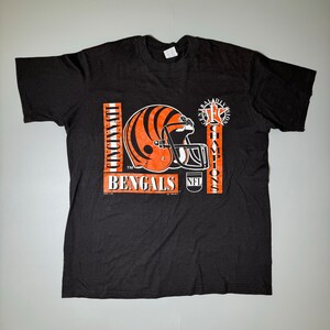 May include: Black t-shirt featuring a Cincinnati Bengals football helmet in orange and black stripes. The shirt includes the text "CINCINNATI BENGALS" and "NFL CENTRAL DIVISION CHAMPIONS" in orange and white.