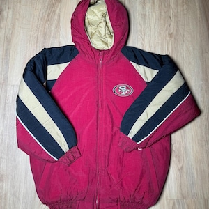 Könnte beinhalten: Rote, schwarze und goldfarbene Kapuzenjacke mit durchgehendem Reißverschluss. Die Jacke hat das Logo der San Francisco 49ers auf der Brust. Die Ärmel haben schwarze, goldene und weiße Streifen. Die Kapuze ist mit einem gesteppten goldenen Material gefüttert.