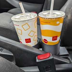 Puede incluir: Dos vasos de refresco de McDonald's con pajitas en el portavasos de un coche. Uno es blanco con formas de diamante amarillas, el otro es amarillo con rayas blancas. Ambos tienen el logotipo de McDonald's.