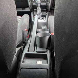 Puede incluir: Portavasos de coche de pl&aacute;stico negro con dos aberturas circulares, colocado entre los asientos delanteros. El interior del coche incluye una palanca de cambios, radio y una moneda en la consola.
