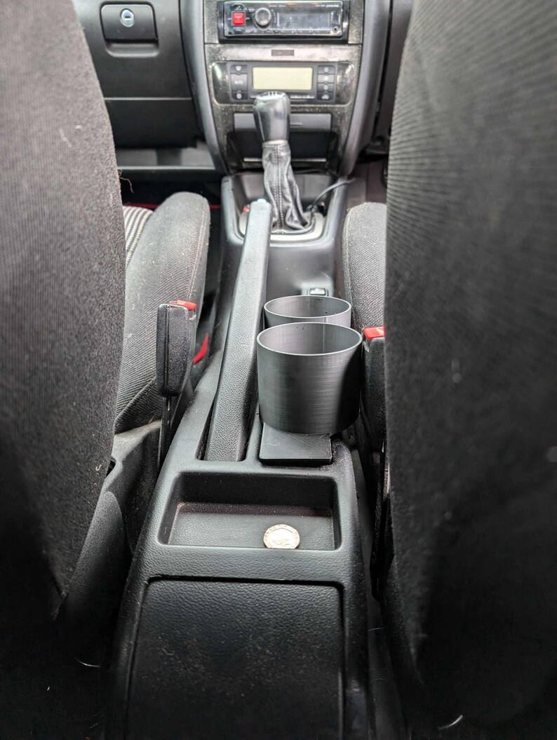 Puede incluir: Portavasos de coche de pl&aacute;stico negro con dos aberturas circulares, colocado entre los asientos delanteros. El interior del coche incluye una palanca de cambios, radio y una moneda en la consola.