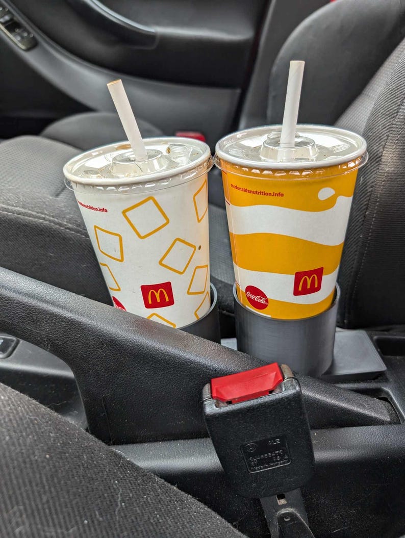 Puede incluir: Dos vasos de refresco de McDonald's con pajitas en el portavasos de un coche. Uno es blanco con cuadrados amarillos y el logo de McDonald's. El otro es amarillo con rayas blancas y el logo de Coca-Cola.