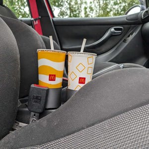 Puede incluir: Dos vasos de refresco de McDonald's con pajitas en un portavasos de coche. Uno tiene un dise&ntilde;o ondulado amarillo y blanco, el otro un fondo blanco con formas de diamante amarillas. Ambos vasos muestran el logotipo de McDonald's.