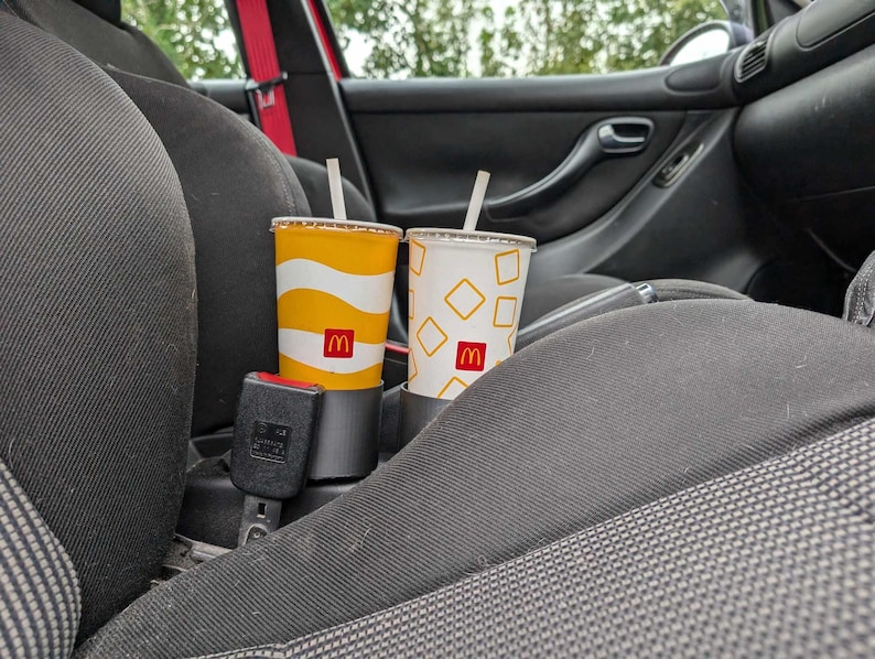 Puede incluir: Dos vasos de refresco de McDonald's con pajitas en un portavasos de coche. Uno tiene un dise&ntilde;o ondulado amarillo y blanco, el otro un fondo blanco con formas de diamante amarillas. Ambos vasos muestran el logotipo de McDonald's.