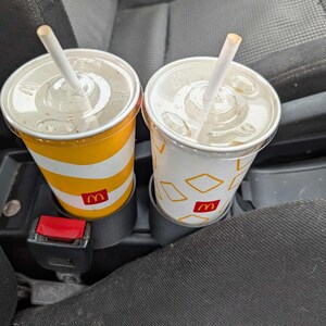 Puede incluir: Dos vasos de refresco de McDonald's con pajitas, uno a rayas amarillas y blancas, el otro blanco con formas de diamante amarillas. Ambos vasos tienen el logotipo de McDonald's y est&aacute;n en los portavasos de un coche.