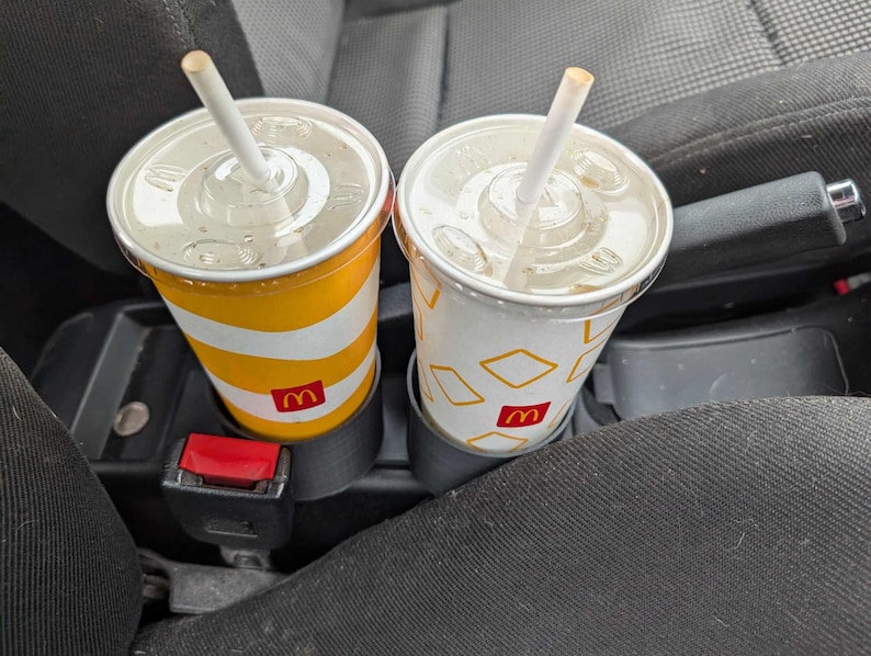 Puede incluir: Dos vasos de refresco de McDonald's con pajitas, uno a rayas amarillas y blancas, el otro blanco con formas de diamante amarillas. Ambos vasos tienen el logotipo de McDonald's y est&aacute;n en los portavasos de un coche.
