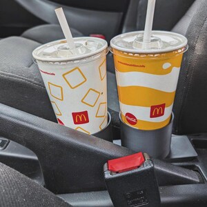 Puede incluir: Dos vasos de refresco de McDonald's con pajitas en los portavasos de un coche. Uno tiene un fondo blanco con cuadrados amarillos y el logotipo de McDonald's. El otro tiene un dise&ntilde;o a rayas amarillas y blancas y el logotipo de Coca-Cola.