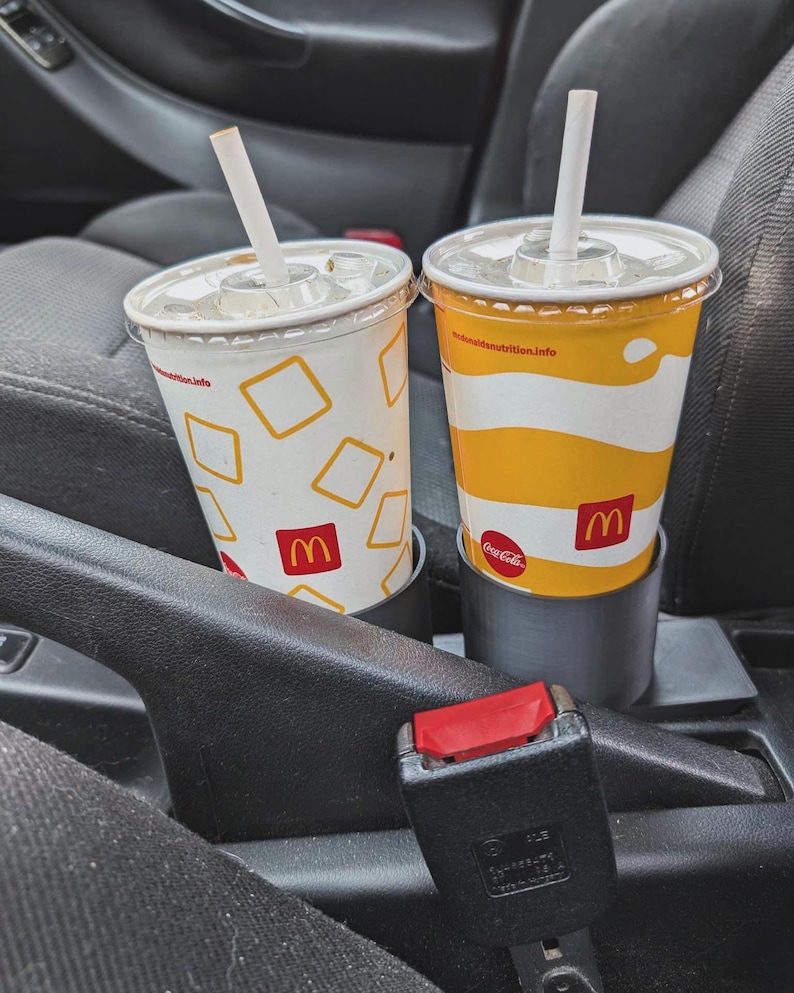 Puede incluir: Dos vasos de refresco de McDonald's con pajitas en los portavasos de un coche. Uno tiene un fondo blanco con cuadrados amarillos y el logotipo de McDonald's. El otro tiene un dise&ntilde;o a rayas amarillas y blancas y el logotipo de Coca-Cola.