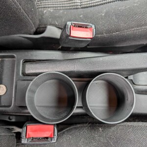 Puede incluir: Portavasos de pl&aacute;stico negro para coche, dise&ntilde;ados para encajar en la consola central de un veh&iacute;culo. Los portavasos son cil&iacute;ndricos con un interior liso y est&aacute;n colocados entre los asientos delanteros.
