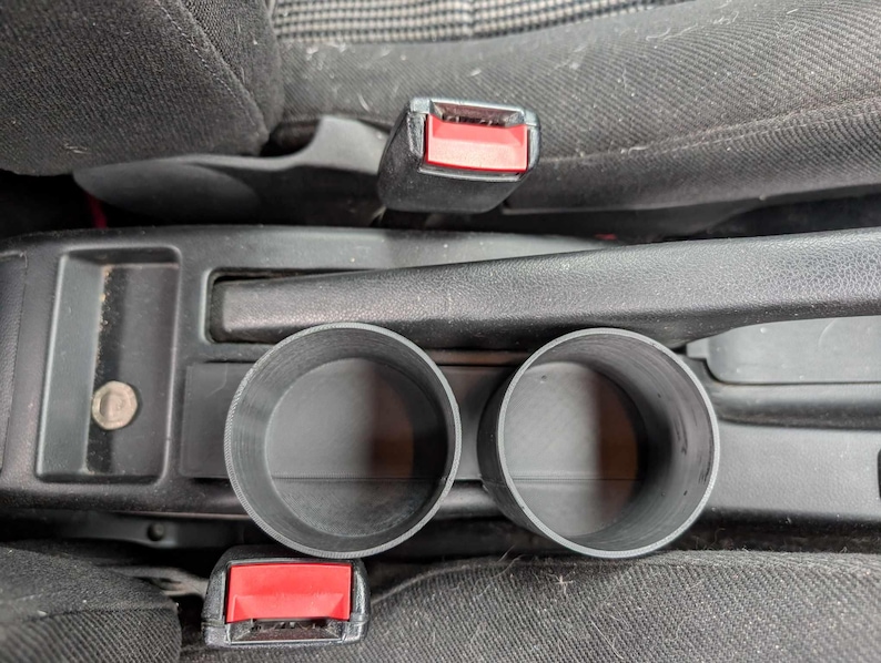 Puede incluir: Portavasos de pl&aacute;stico negro para coche, dise&ntilde;ados para encajar en la consola central de un veh&iacute;culo. Los portavasos son cil&iacute;ndricos con un interior liso y est&aacute;n colocados entre los asientos delanteros.