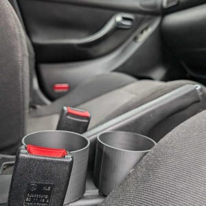 Puede incluir: Interior de coche con una hebilla de cintur&oacute;n de seguridad negra con un bot&oacute;n de liberaci&oacute;n rojo y dos portavasos grises. El interior del coche es negro con asientos de tela gris.