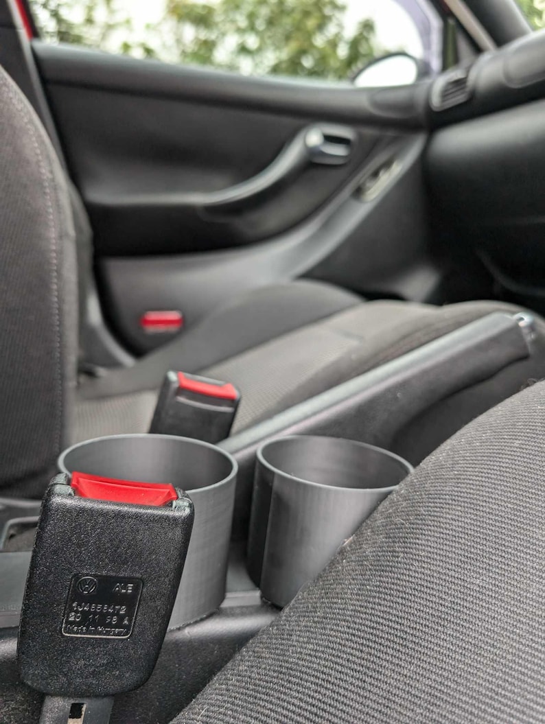 Puede incluir: Interior de coche con una hebilla de cintur&oacute;n de seguridad negra con un bot&oacute;n de liberaci&oacute;n rojo y dos portavasos grises. El interior del coche es negro con asientos de tela gris.