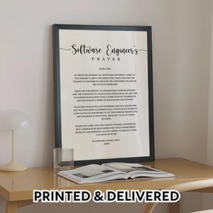 May include: A framed print with the text "Software Engineer's Prayer" in elegant script. The black frame surrounds the text, which is centered on a white background. A glass and an open book are on the table. The text "PRINTED & DELIVERED" is at the bottom.
