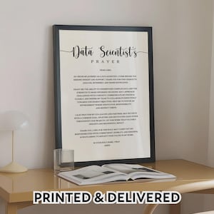 May include: A framed print featuring the "Data Scientist's Prayer" in a stylish script. The black frame surrounds the white paper with the prayer text. A glass of water and an open book are on the table below. The words "PRINTED & DELIVERED" are at the bottom.