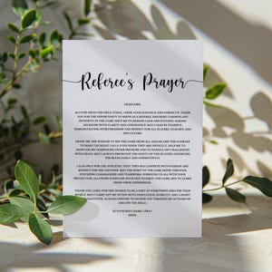May include: A white card with the text "Referee's Prayer" in elegant script. The card contains a prayer for referees, with text in a classic serif font. Green foliage frames the card, adding a natural touch.