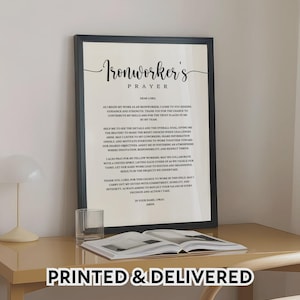 May include: A framed print with the text "Ironworker's Prayer" in elegant script. The print is displayed on a light-colored desk, alongside a glass and an open book. The phrase "PRINTED & DELIVERED" is at the bottom.
