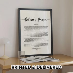 Könnte beinhalten: Ein gerahmter Druck mit dem Text "Referee's Prayer" in eleganter Schrift. Der schwarze Rahmen umgibt einen weißen Hintergrund mit dem Text des Gebets in einer klassischen Schriftart. Der Druck wird auf einem hellen Schreibtisch mit einem Buch und einem Glas präsentiert.