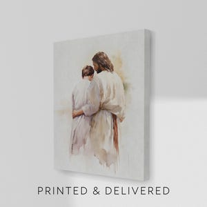 May include: A canvas print featuring a watercolor painting of two figures embracing. One figure, with long brown hair, is in a white robe, embracing another figure in a light purple robe. The text "PRINTED & DELIVERED" is at the bottom.