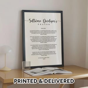 May include: A framed print featuring the "Software Developer's Prayer" in a stylish script. The black frame encloses the text on a white backdrop. A glass of water and an open book are on the table. The words "PRINTED & DELIVERED" are at the bottom of the image.