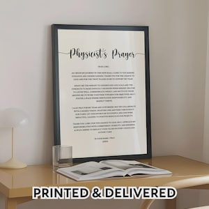 May include: A framed print with the text "Physicist's Prayer" in elegant script. The black frame surrounds a white background with the prayer text. A glass and an open book are on the table below. The words "PRINTED & DELIVERED" are at the bottom.