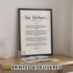 May include: A framed print with the text "App Developer's Prayer" in elegant script. The black frame surrounds a white background with the prayer text. The words "PRINTED & DELIVERED" are at the bottom. A glass and open book are on the table.