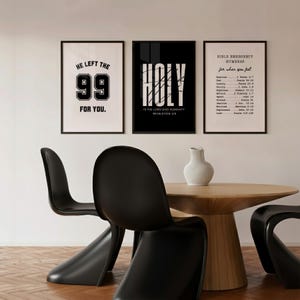 Puede incluir: Tres impresiones artísticas enmarcadas con marcos negros. Las impresiones presentan el texto: "HE LEFT THE 99 FOR YOU.", "HOLY IS THE LORD GOD ALMIGHTY REVELATION 4:3" y "BIBLE EMERGENCY NUMBERS for when you feel". Las impresiones se muestran sobre una mesa de madera y sillas negras.