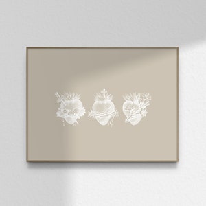 May include: A beige framed print featuring three white illustrations of stylised hearts with thorns, flowers, and a sword. The hearts are arranged horizontally.