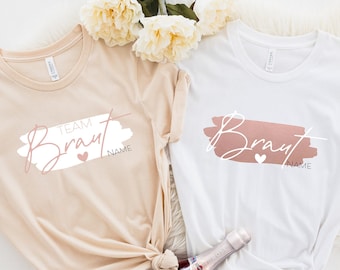 Könnte beinhalten: Zwei weiße T-Shirts mit einem rosa und weißen Design, auf dem "Team Braut" und "Braut" mit einem Herz steht. Die Shirts liegen auf einem weißen Hintergrund mit einer Flasche Champagner.