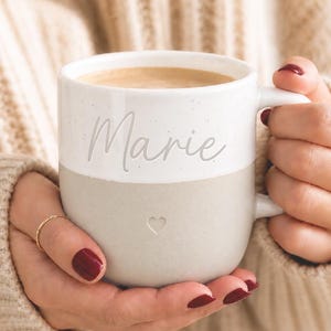 Könnte beinhalten: Ein zweifarbiger Keramikbecher mit einem weiß gesprenkelten oberen Teil, auf dem der Name Marie in Schreibschrift steht, und einem einfarbigen hellbeigen unteren Teil. Ein kleines, schlichtes Herz ist in der Mitte des beigen Bereichs eingeprägt.