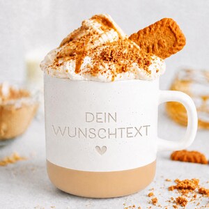 Könnte beinhalten: Ein weißer Keramikbecher mit beigefarbenem Boden, der den geprägten Text DEIN WUNSCHTEXT und ein kleines Herzsymbol aufweist. Der Becher ist mit Schlagsahne gefüllt, garniert mit Karamellsauce, Keksbröseln und einem einzelnen rechteckigen Keks.